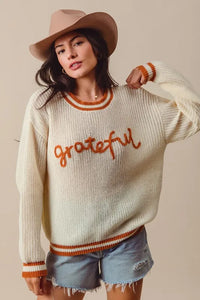 Woman wearing a sweater with 'grateful' text, denim shorts, and a hat on a beige background