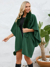 Woman wearing a green poncho with fur trim against a white wall.
