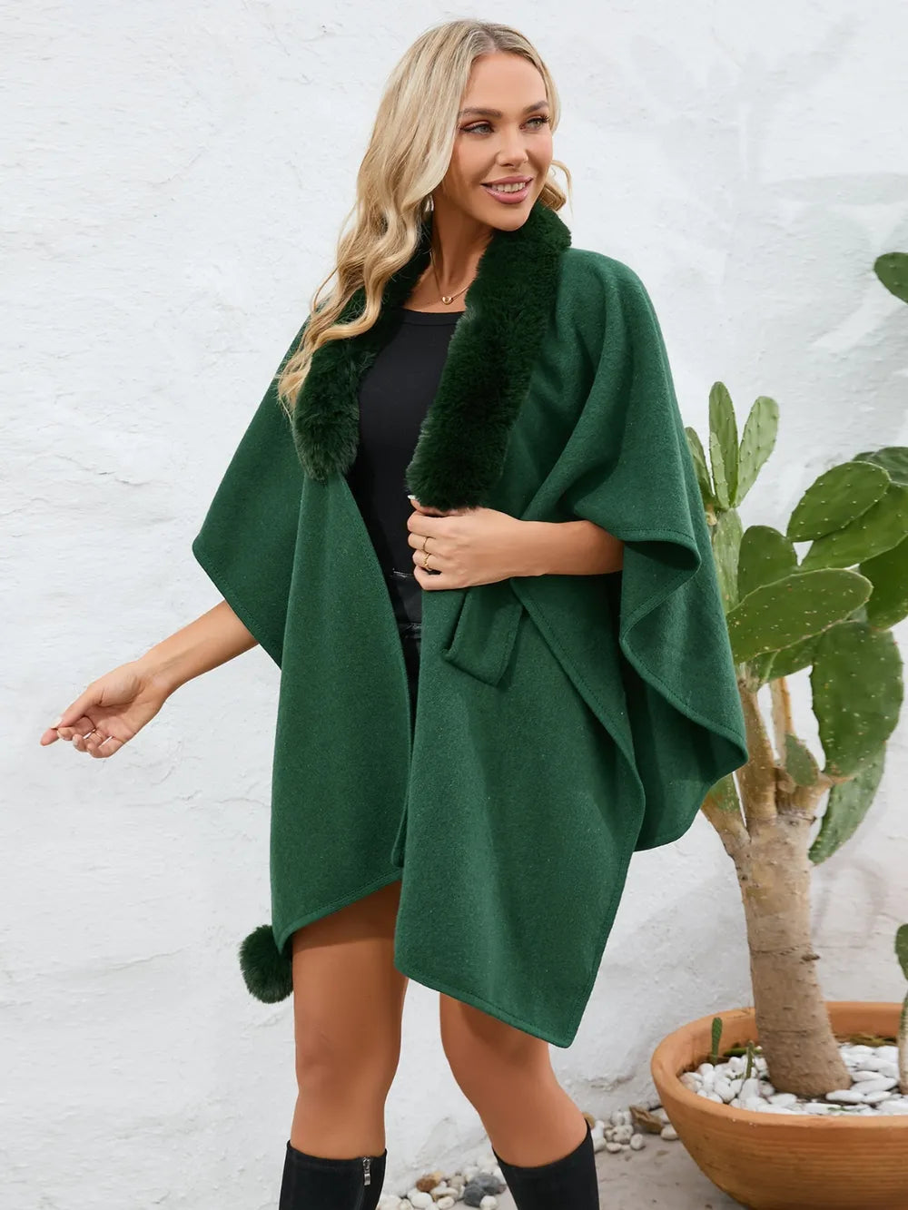 Woman wearing a green poncho with fur trim against a white wall.