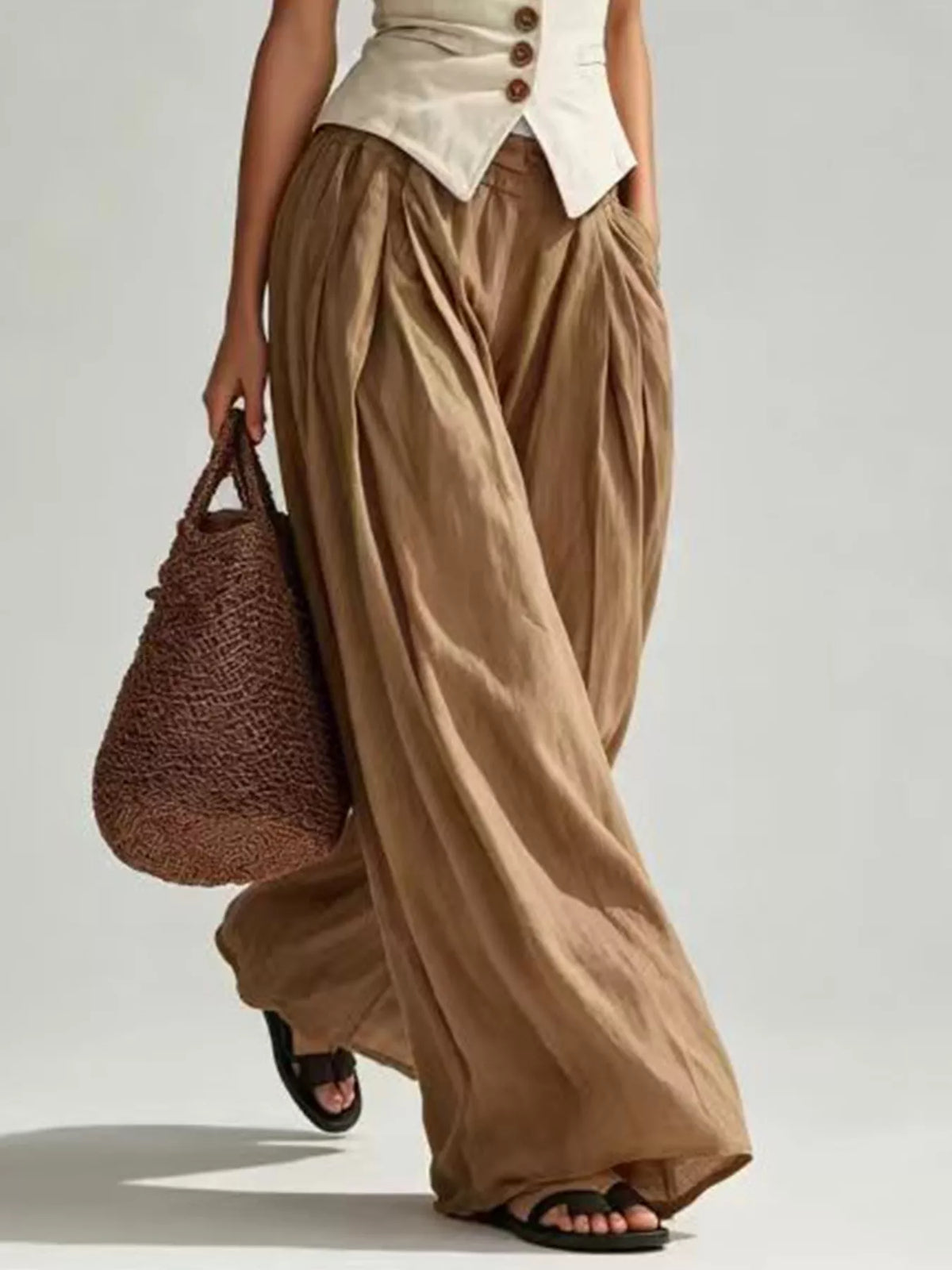 A model wearing voluminous, draped wide-leg pants in a warm taupe-brown color, styled with a cream button-up top, black slide sandals, and carrying a brown woven tote bag against a light gray studio background.