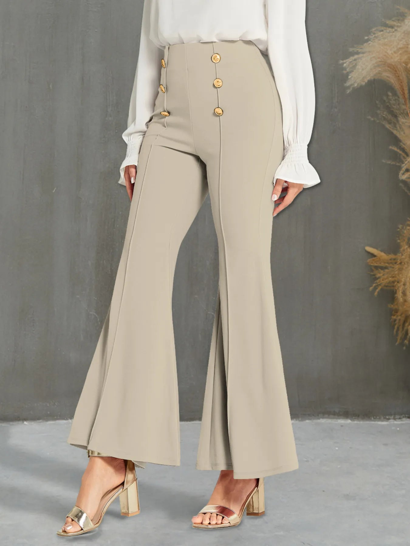 Beige flared pants with gold buttons worn by a person against a gray background