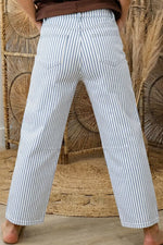 Person wearing white and blue striped pants standing against a woven wall.