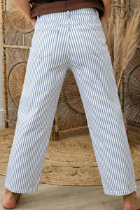 Person wearing white and blue striped pants standing against a woven wall.