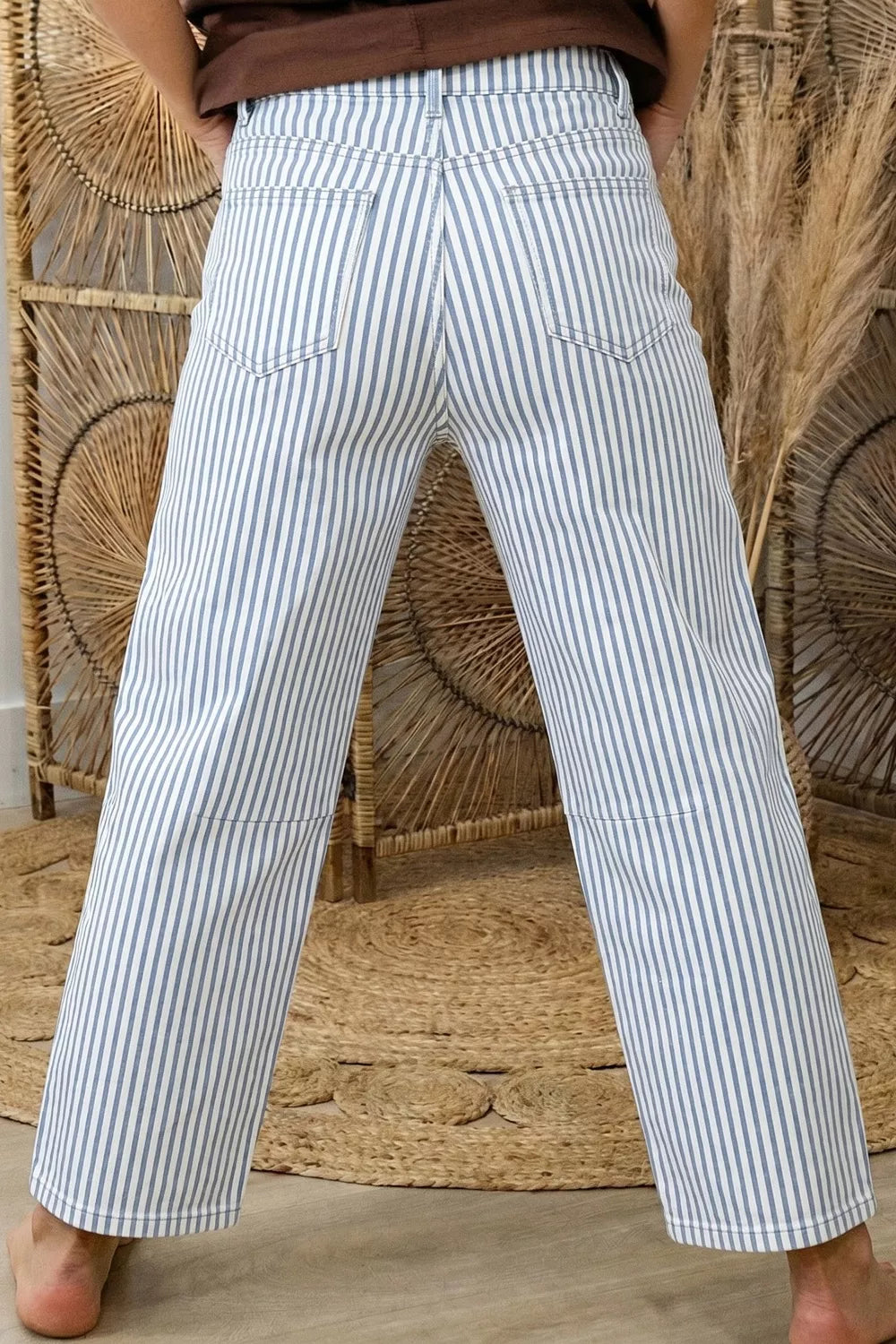 Person wearing white and blue striped pants standing against a woven wall.