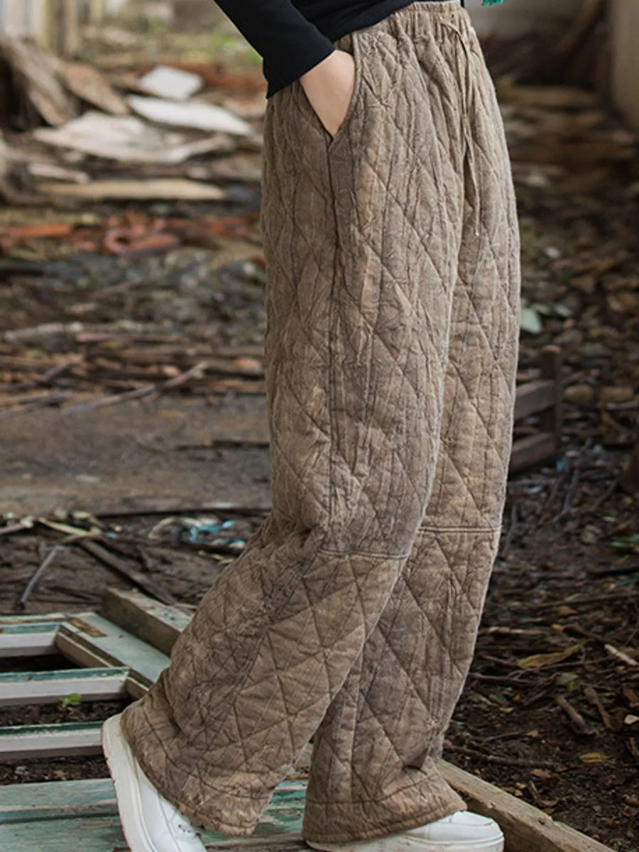 A person wearing loose, quilted wide-leg pants in a taupe-brown color, standing outdoors on a weathered wooden platform with a messy, debris-filled background.