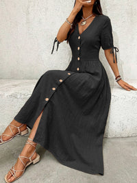 Woman wearing a black dress with button details sitting against a light gray wall.
