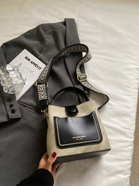 Handbag with black flap and beige body on a bed with a gray jacket and card.
