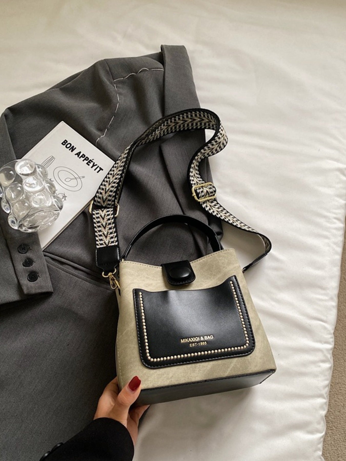 Handbag with black flap and beige body on a bed with a gray jacket and card.
