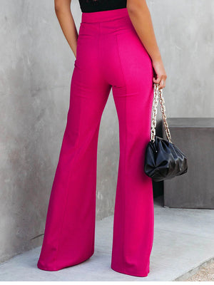 Person wearing bright pink pants holding a black handbag against a gray wall.