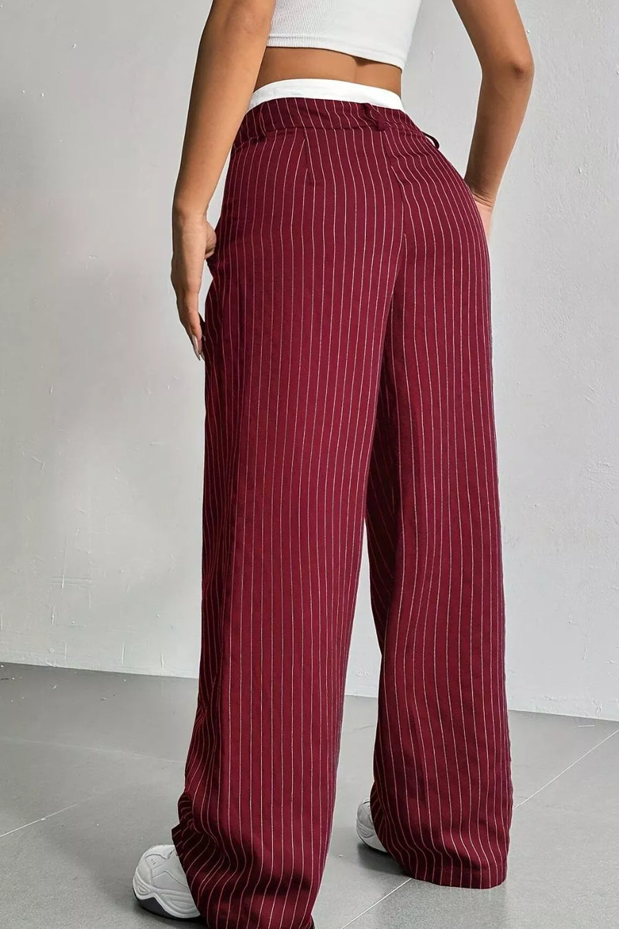 A person wearing high-waisted, wide-leg trousers in burgundy with thin white vertical pinstripes, shown from the back against a light gray wall and floor.