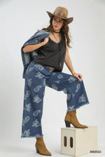 Woman wearing a black top, blue paisley pants, and brown cowboy hat on a white background
