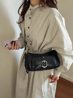 A person wearing an oversized light beige shirt dress carries a black faux-leather shoulder bag with a silver buckle. The person is holding a light purple phone in a home setting with a beige wall and wooden floor.