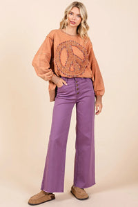 Woman wearing a brown peace sign shirt and purple pants against a beige background