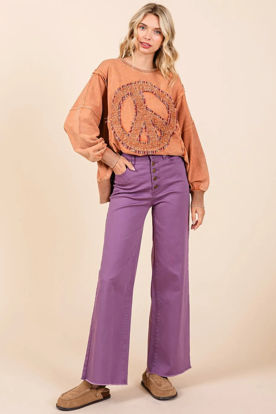 Woman wearing a brown peace sign shirt and purple pants against a beige background