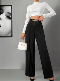 Person wearing a white long-sleeve top and black high-waisted pants, holding a white handbag against a gray background.