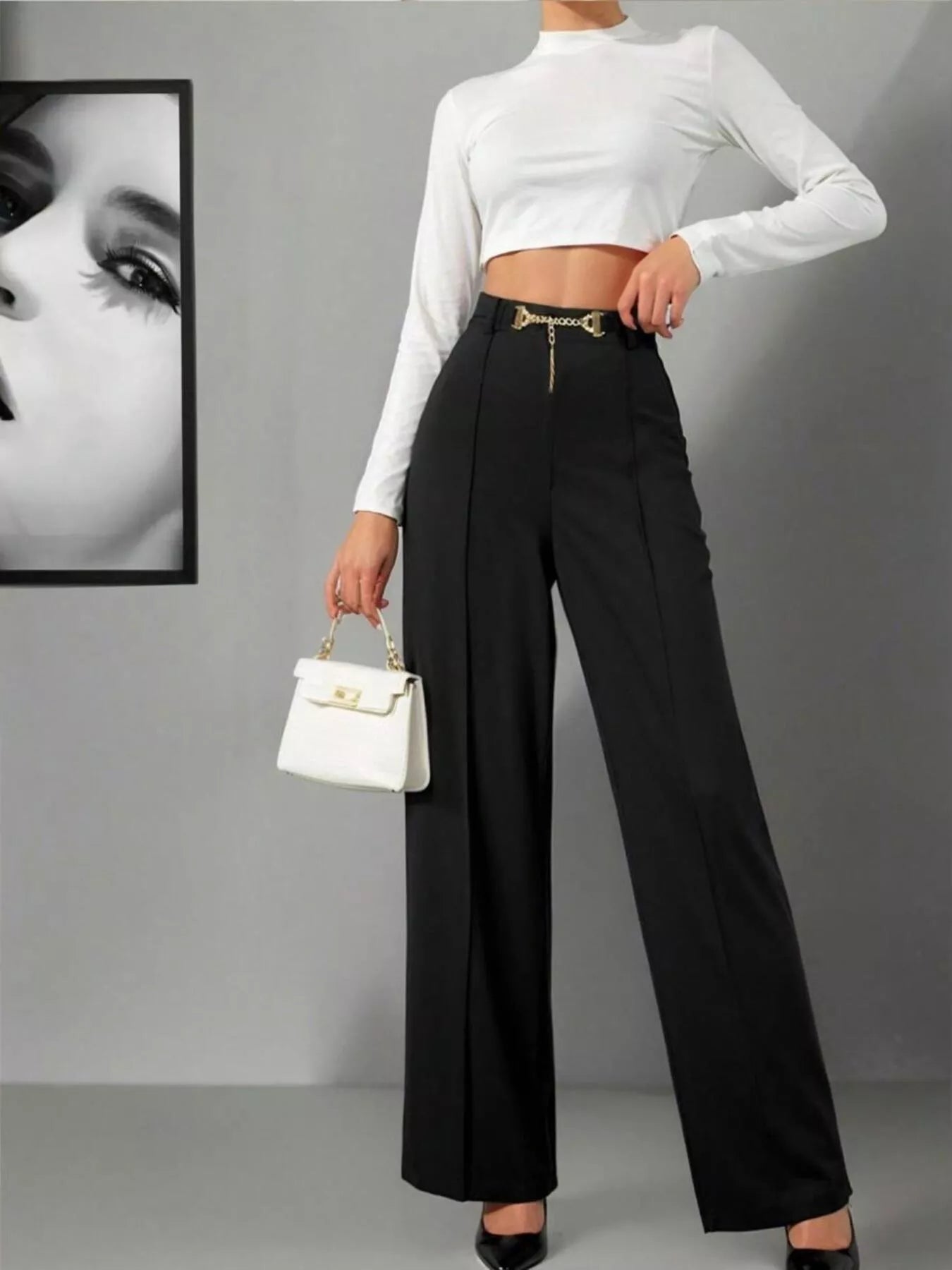 Person wearing a white long-sleeve top and black high-waisted pants, holding a white handbag against a gray background.
