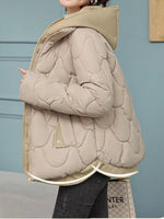 A person wearing a light beige quilted puffer jacket with a hood, paired with dark gray jeans, standing against a muted blue-gray paneled wall.