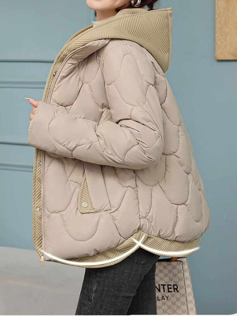 A person wearing a light beige quilted puffer jacket with a hood, paired with dark gray jeans, standing against a muted blue-gray paneled wall.