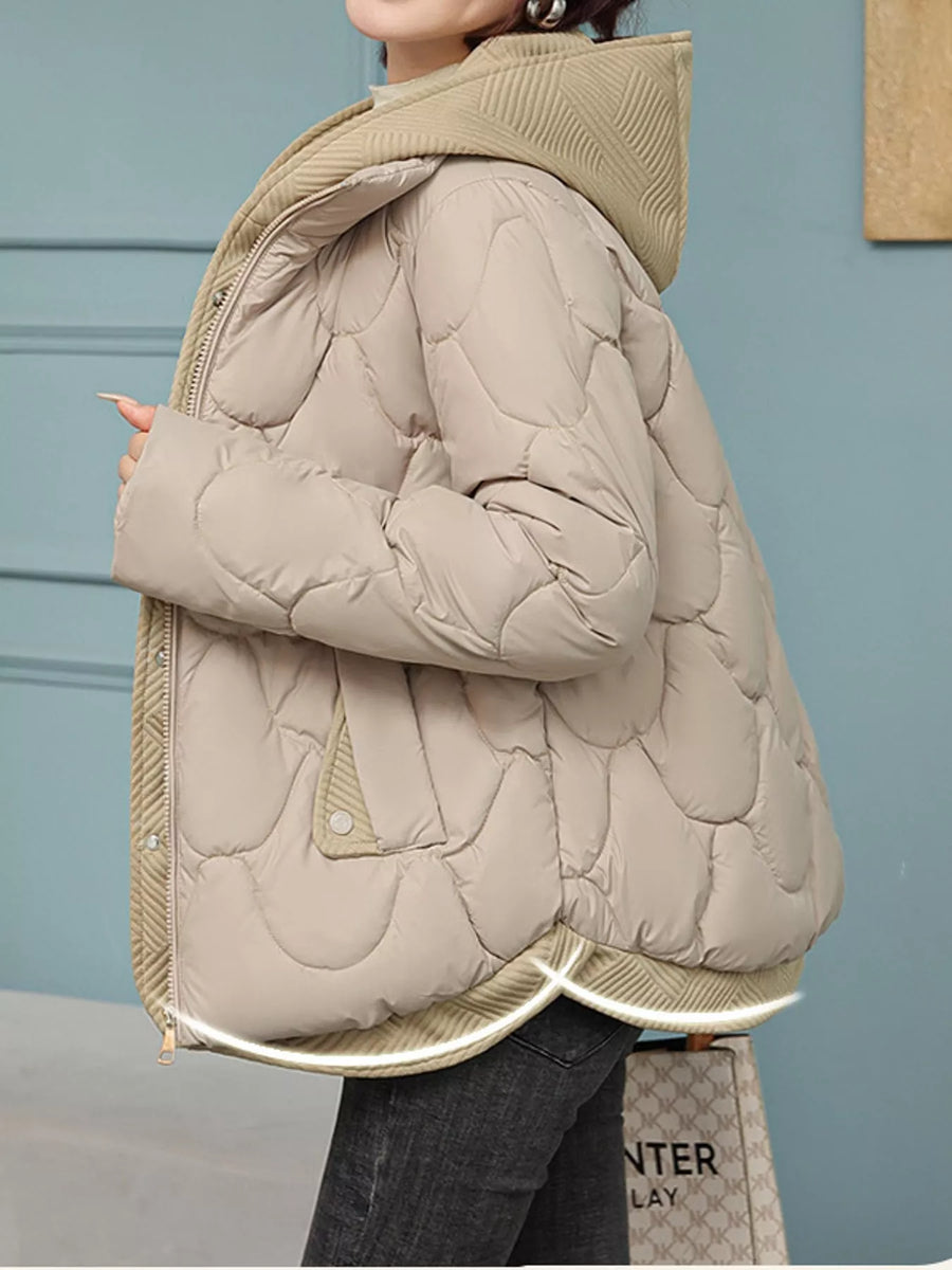 A person wearing a light beige quilted puffer jacket with a hood, paired with dark gray jeans, standing against a muted blue-gray paneled wall.