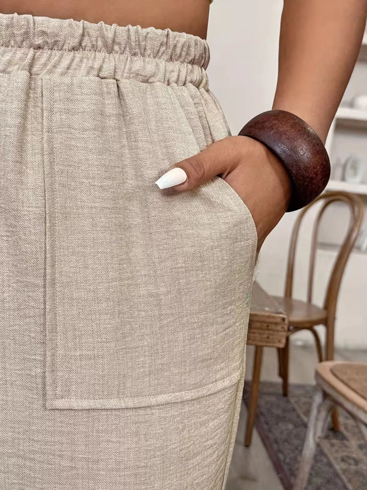 Close-up of a person wearing a light beige linen-textured skirt with an elastic waistband and a large front patch pocket, with one hand tucked into the pocket. The person is wearing a thick dark brown wooden cuff bracelet.