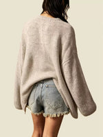 A person shown from the back wearing a loose, oversized ribbed knit sweater in a light warm gray-beige tone, paired with light-wash denim shorts with frayed hems, against a plain light beige background.