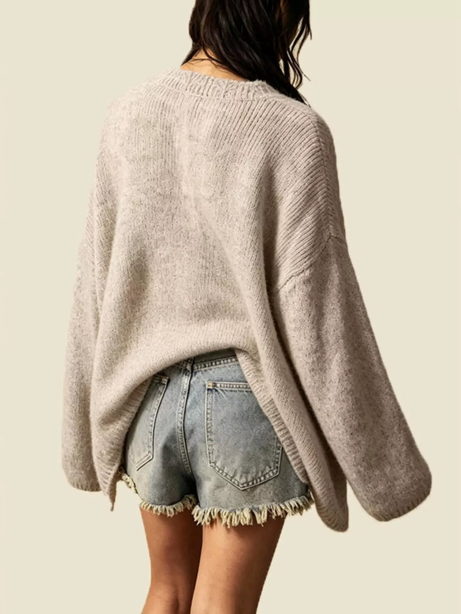 A person shown from the back wearing a loose, oversized ribbed knit sweater in a light warm gray-beige tone, paired with light-wash denim shorts with frayed hems, against a plain light beige background.