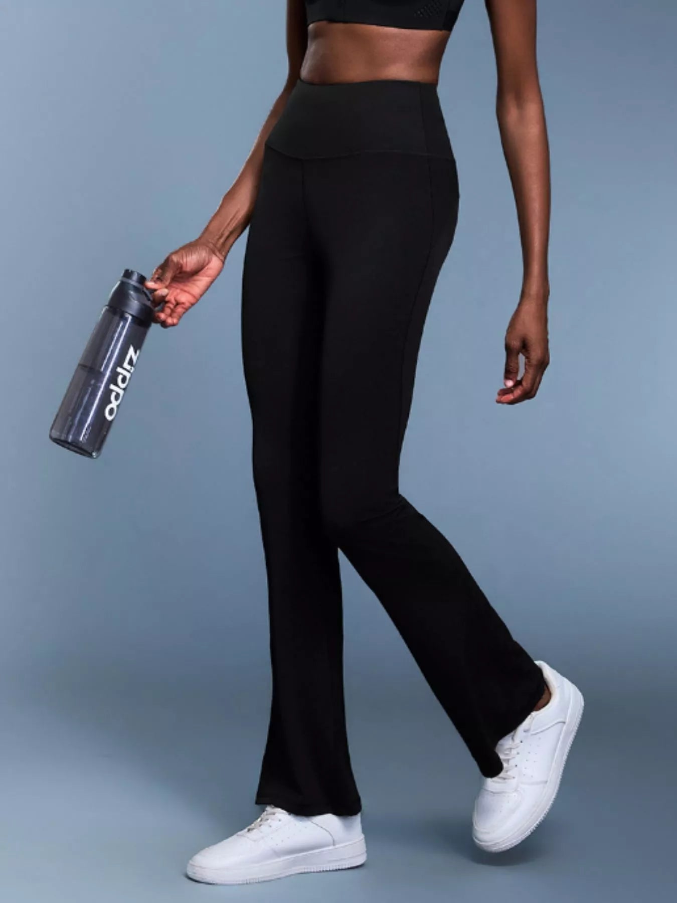 Person wearing black leggings and white sneakers holding a black water bottle with a brand logo on a gray background