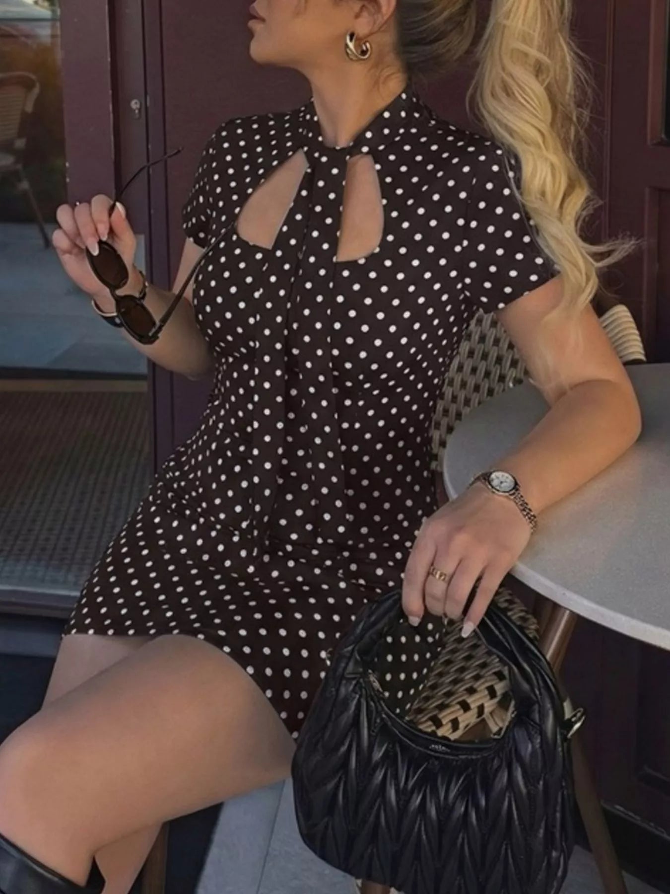 Woman in a black polka dot dress holding sunglasses and a black handbag.