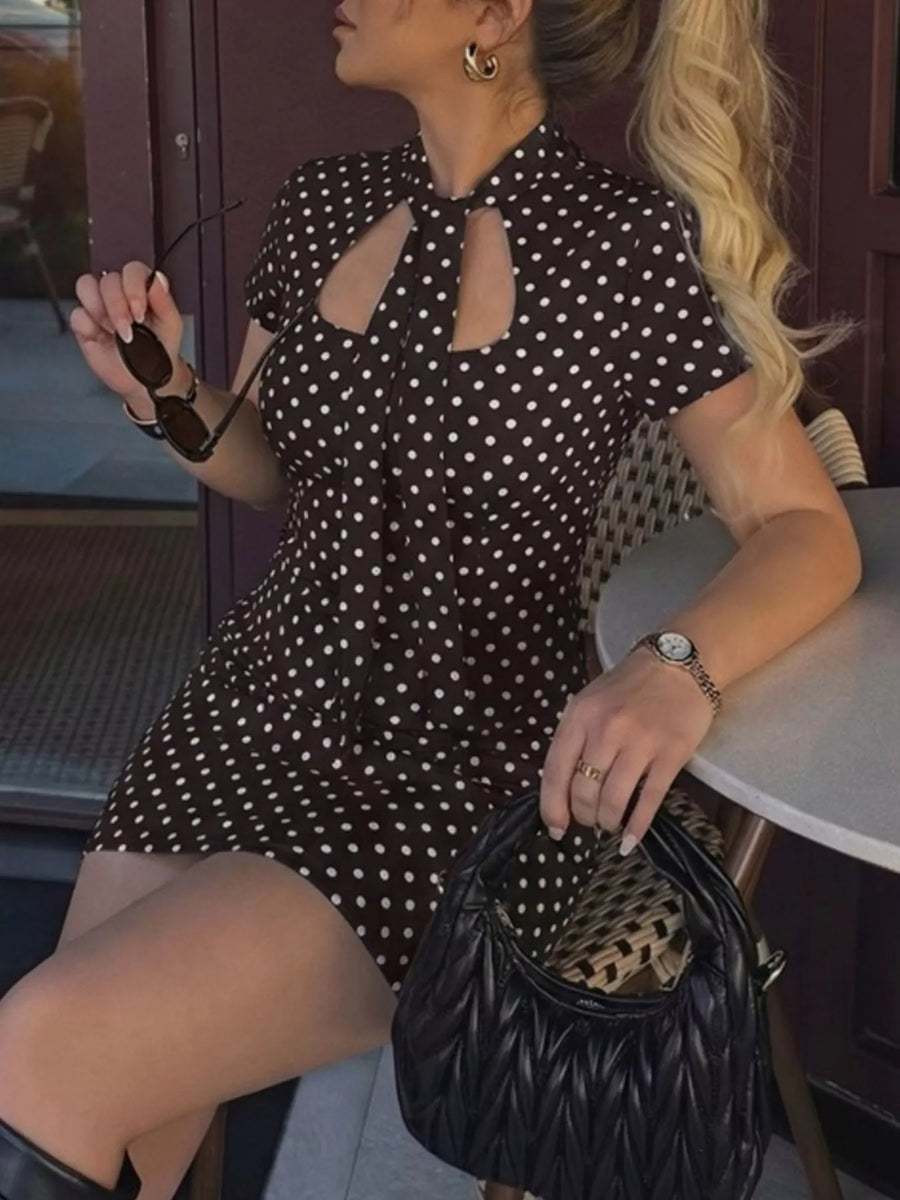 Woman in a black polka dot dress holding sunglasses and a black handbag.