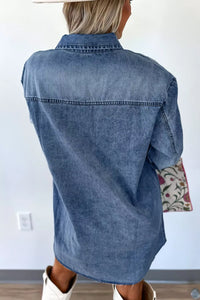 Person wearing a blue denim shirt with rolled-up sleeves, standing against a white wall.