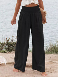 Person wearing black wide-leg pants standing on a sandy beach with a light blue wall in the background.