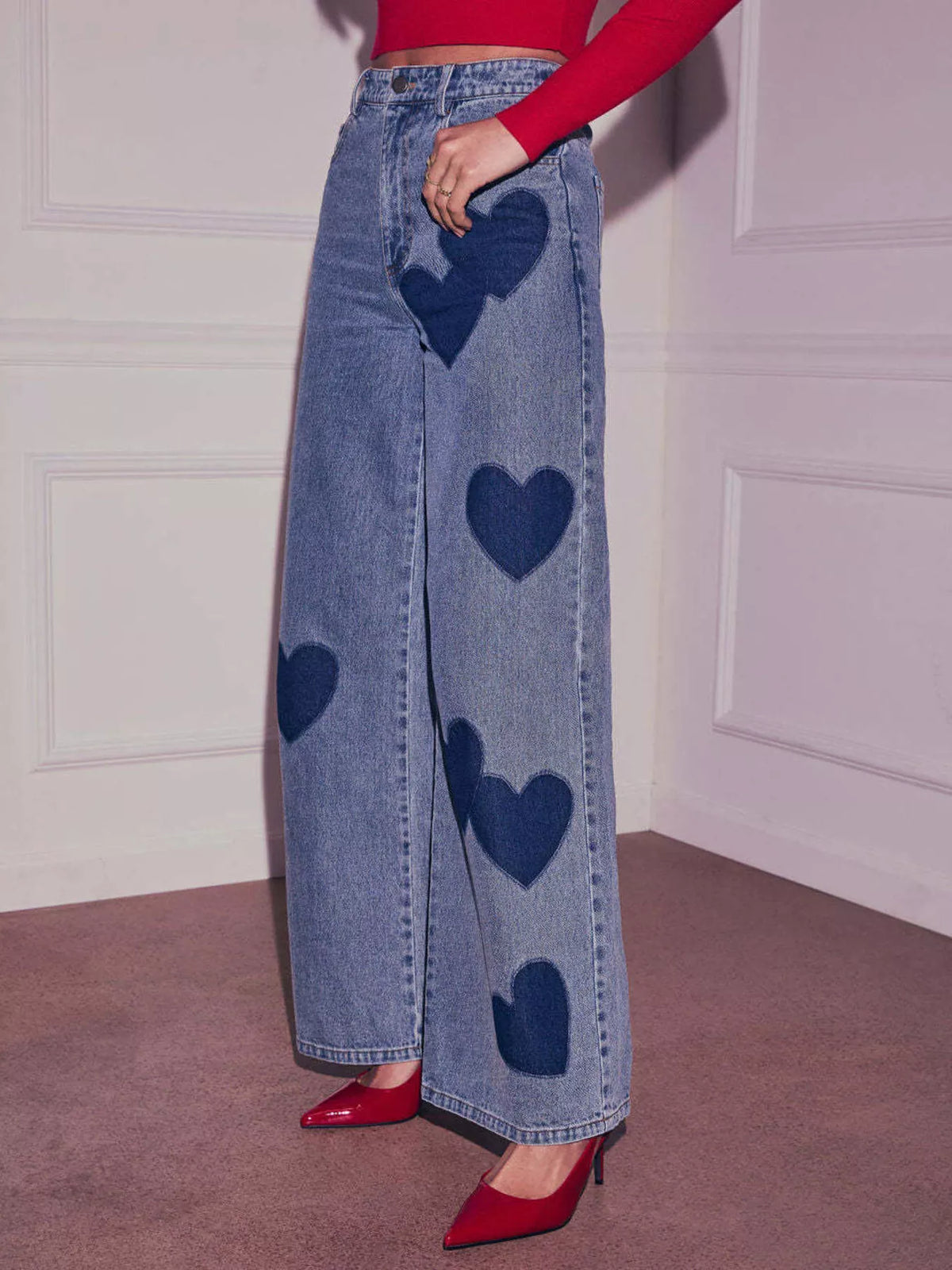 Person wearing blue jeans with heart patterns and red shoes against a white wall.