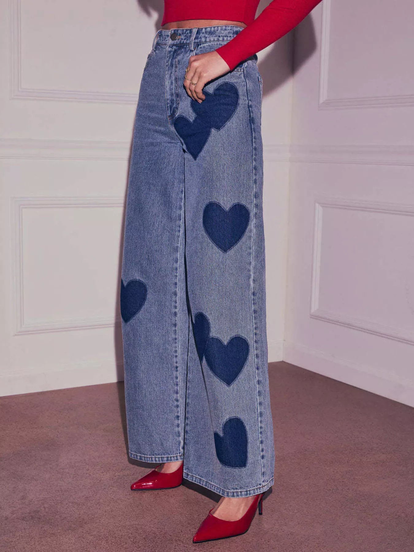 Person wearing blue jeans with heart patterns and red shoes against a white wall.