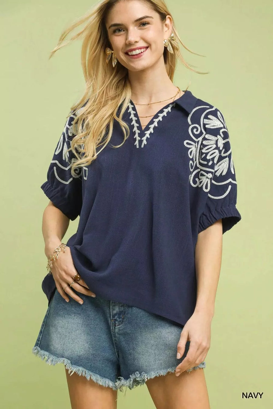 Woman wearing a navy blue blouse with white embroidery and denim shorts on a light green background