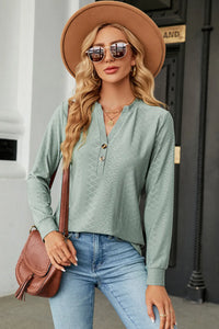 Woman wearing a green long-sleeve top, blue jeans, and a brown hat with sunglasses outdoors.