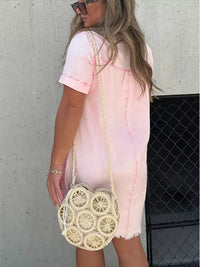 Woman wearing a pink tie-dye dress holding a round woven bag against a gray wall.