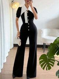 Person wearing a black and white outfit with a high collar, standing in a room with a plant and chair.