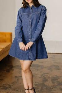 Woman wearing a blue denim dress standing in a room with a wooden floor and a couch.
