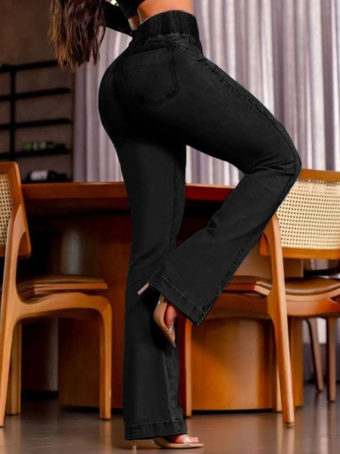 Person wearing black flared jeans sitting on a wooden chair indoors.