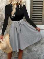 Woman wearing a black long-sleeve top and gray striped skirt, holding a straw hat, against a stone wall.