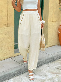 Woman wearing beige high-waisted pants with button details, white top, and sandals, standing against a stone wall.