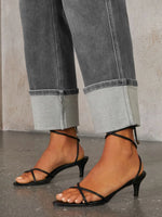 Black strappy high-heeled sandals worn with rolled-up gray jeans on a neutral background