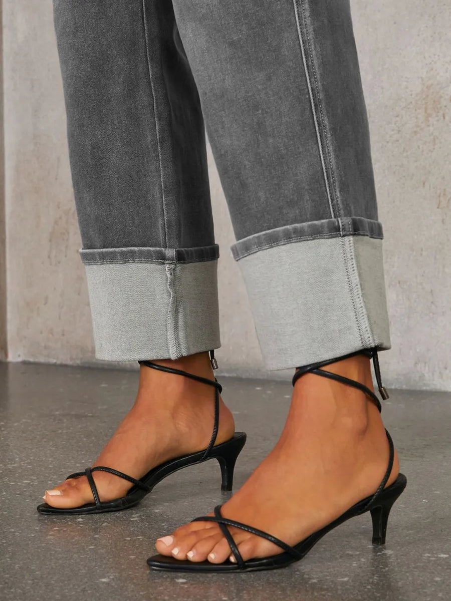 Black strappy high-heeled sandals worn with rolled-up gray jeans on a neutral background