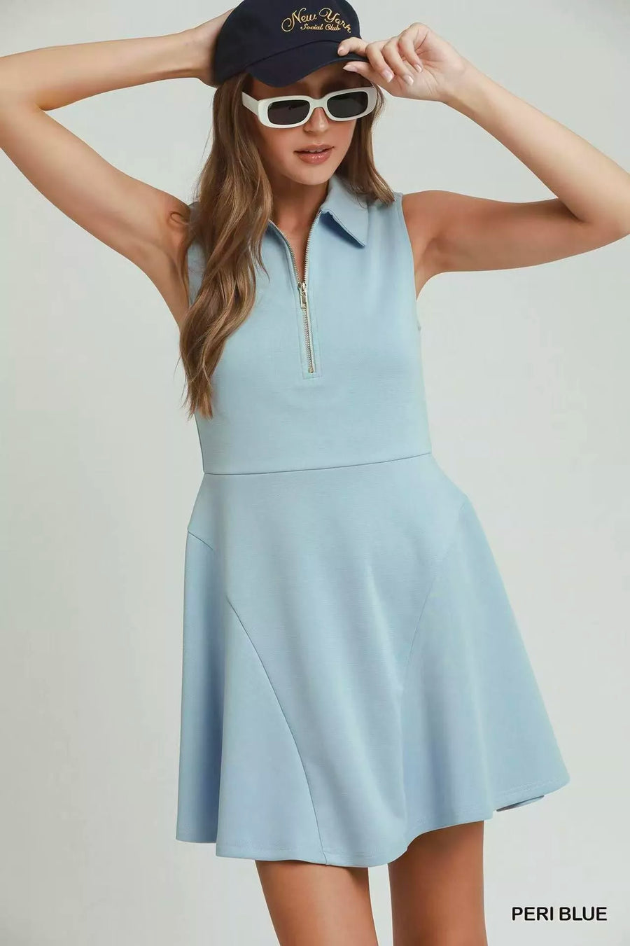 Woman wearing a light blue sleeveless dress with a zipper, black cap, and sunglasses on a plain background.