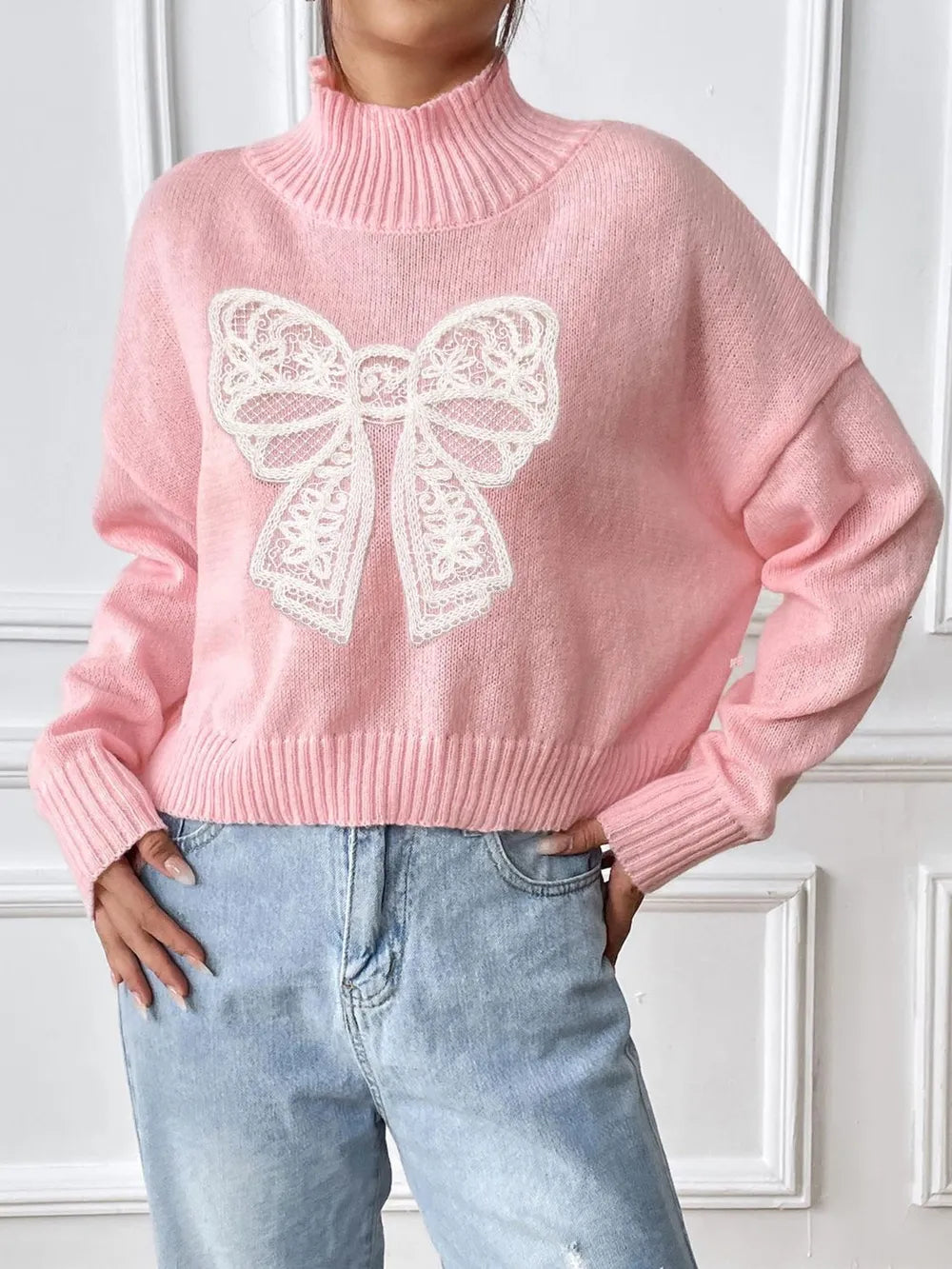 Pink sweater with a large white bow design worn by a person against a white wall.