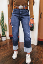 Person wearing blue jeans with a belt buckle, brown shirt, and white sneakers indoors.