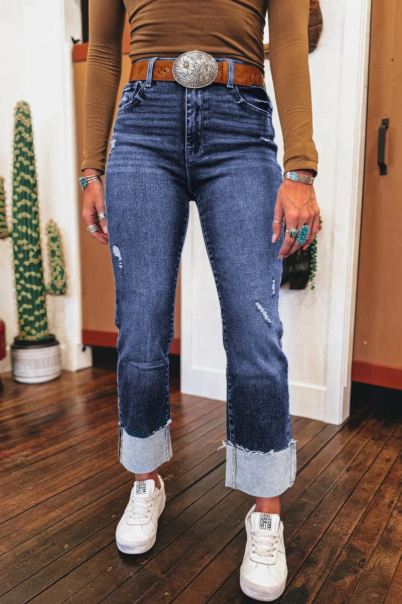 Person wearing blue jeans with a belt buckle, brown shirt, and white sneakers indoors.