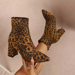A pair of leopard-print ankle boots with pointed toes and block heels, shown being worn with one boot lifted on a foot and the other held in a hand with rings.