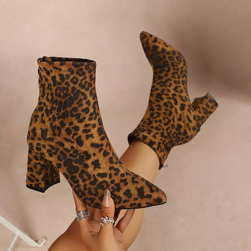 A pair of leopard-print ankle boots with pointed toes and block heels, shown being worn with one boot lifted on a foot and the other held in a hand with rings.