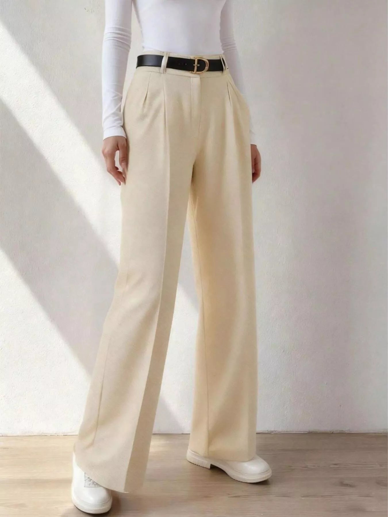 Person wearing beige wide-leg pants with a white shirt and black belt against a plain background
